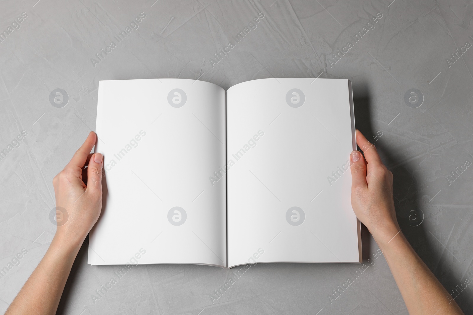 Photo of Woman holding open notebook with blank pages at light grey table, top view. Mockup for design