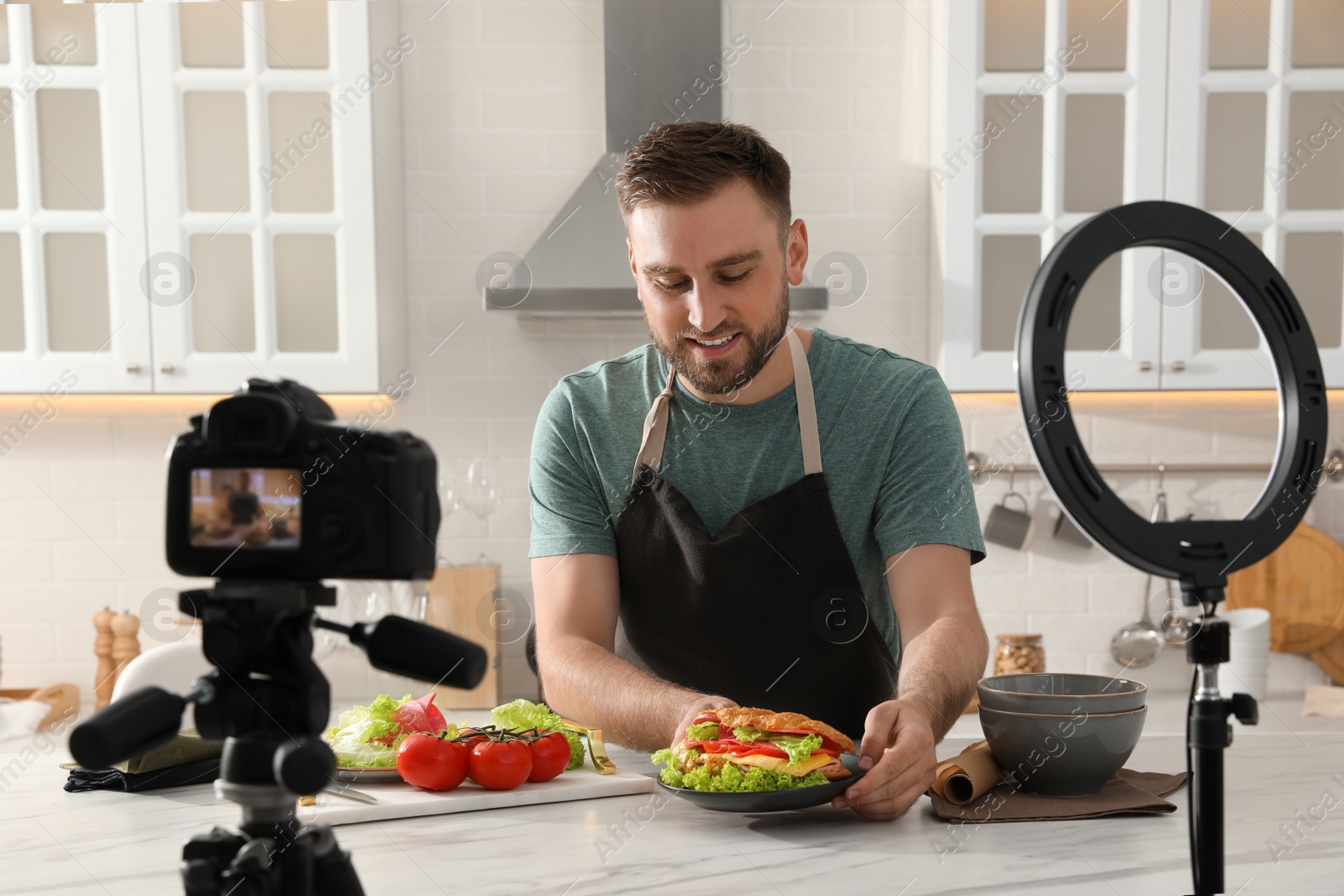 Photo of Blogger with tasty croissant recording video in kitchen at home. Using ring lamp and camera