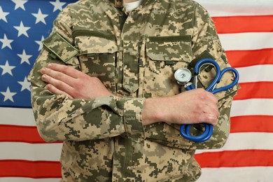 Man in military uniform with crossed arms and stethoscope against USA flag, closeup. Health care concept