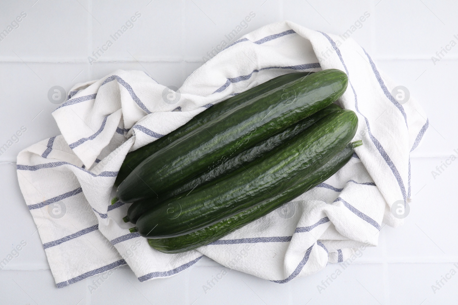 Photo of Fresh cucumbers and cloth on white tiled table, top view