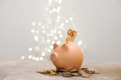 Photo of Piggy bank with euro banknote and coins on grey table against blurred lights, space for text