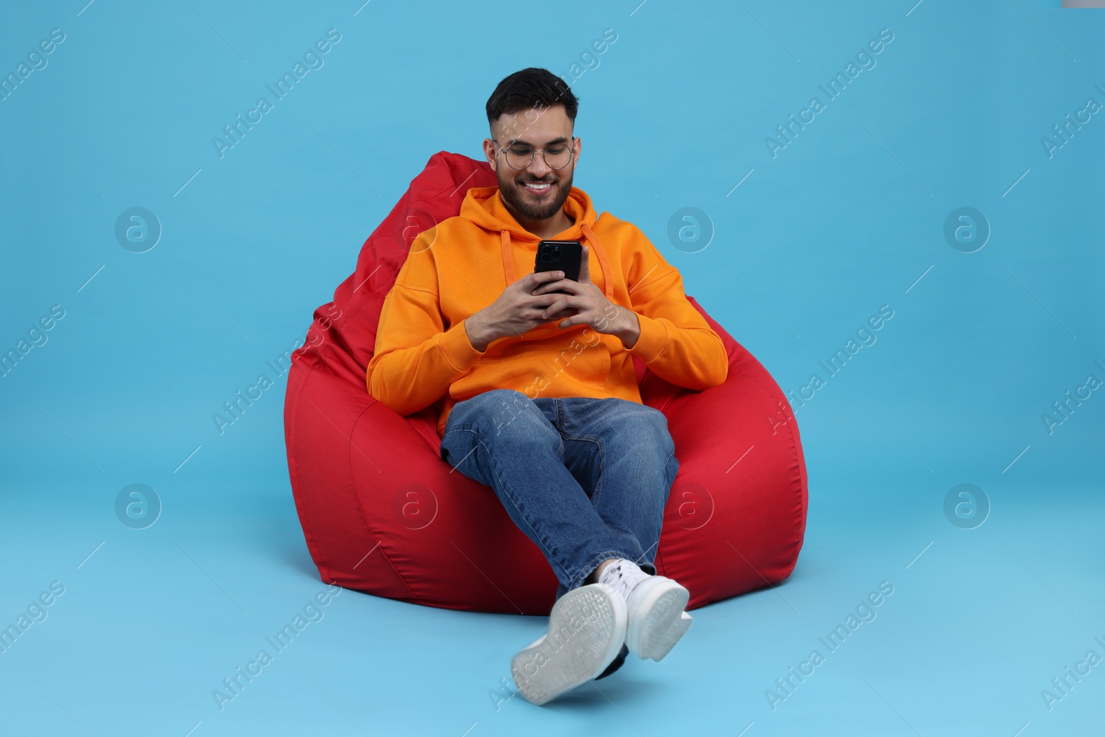 Photo of Happy young man using smartphone on bean bag chair against light blue background