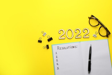 Making New Year's resolutions. Notebook, glasses, clips and number 2022 on yellow background, flat lay with space for text