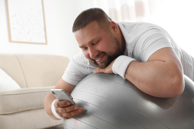 Photo of Lazy overweight man using smartphone while lying on exercise ball at home