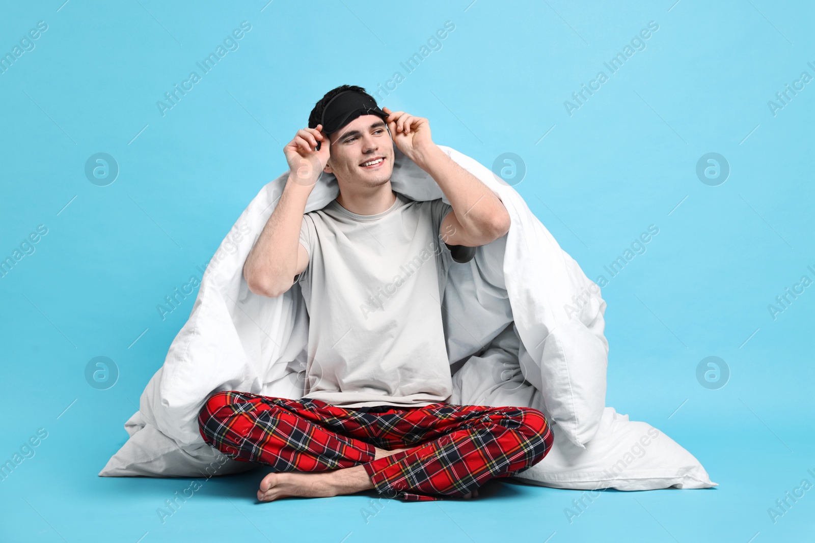 Photo of Happy man in pyjama and sleep mask wrapped in blanket on light blue background