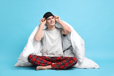 Photo of Happy man in pyjama and sleep mask wrapped in blanket on light blue background