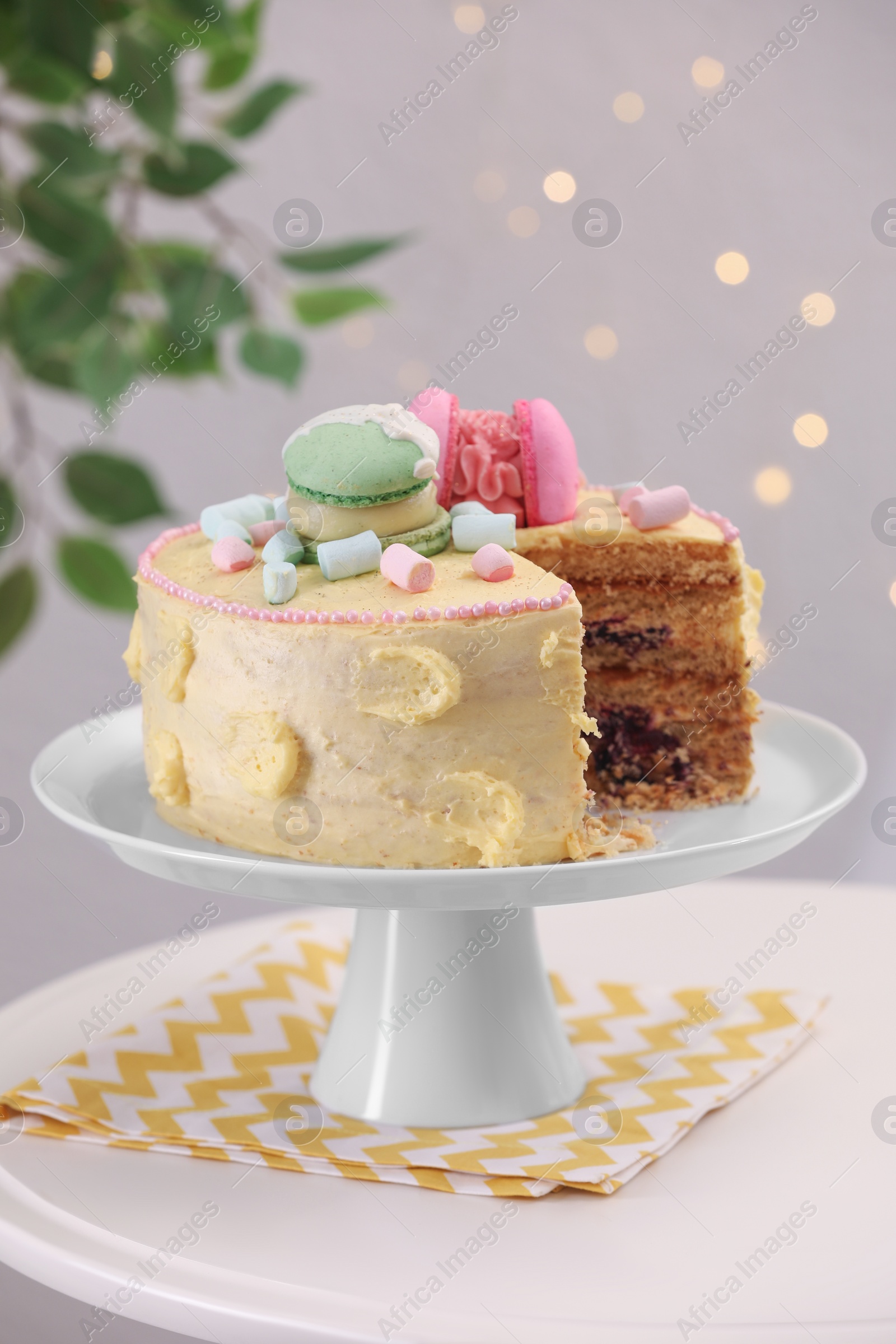 Photo of Delicious cake decorated with macarons and marshmallows on white table against blurred lights