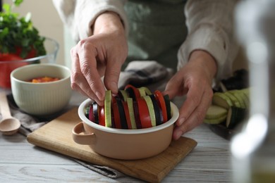 Photo of Cooking delicious ratatouille. Woman adding slice of zuccini into bowl with fresh vegetables at white wooden table, closeup