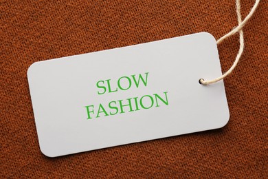 Image of Conscious consumption. Tag with words Slow Fashion on brown fabric, top view
