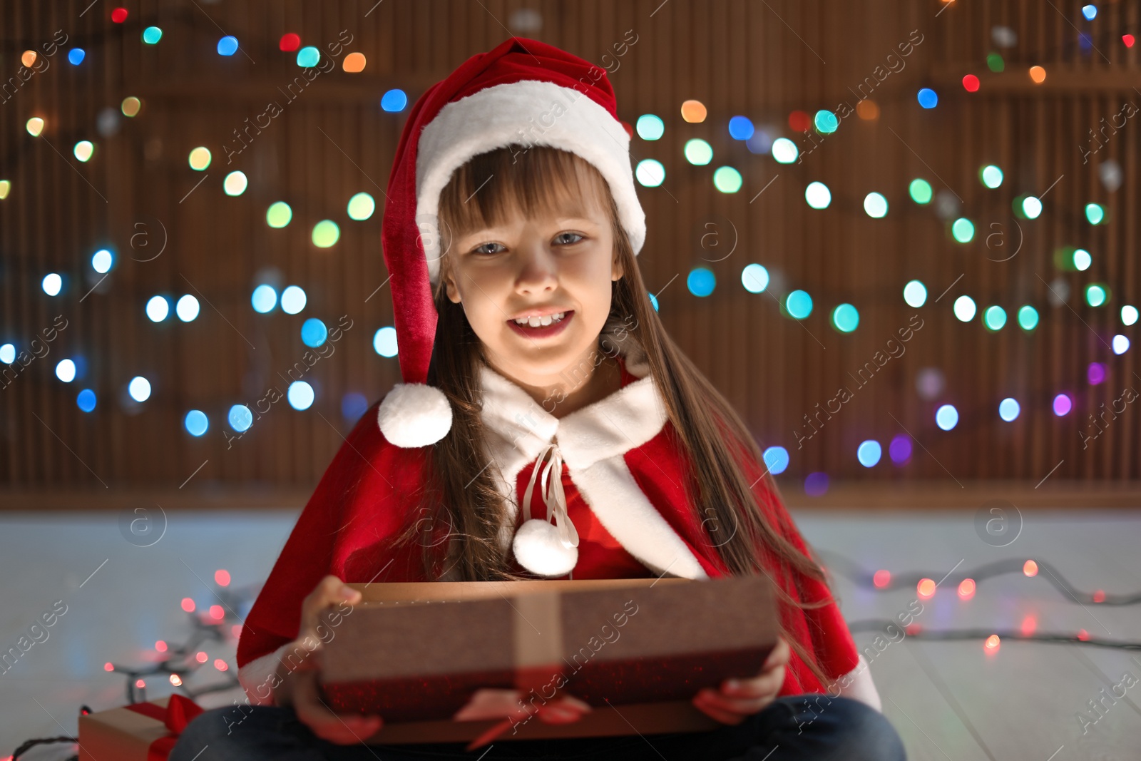 Photo of Cute little child in Santa hat opening Christmas gift on blurred lights background