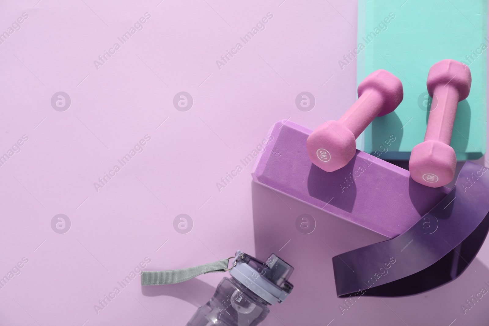 Photo of Two dumbbells, yoga blocks, fitness elastic band and bottle on violet background, flat lay. Space for text