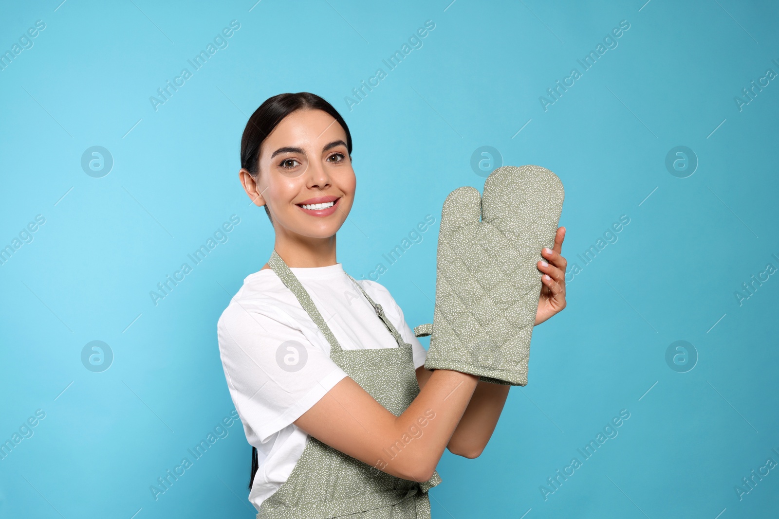 Photo of Young woman in grey apron and oven glove on light blue background