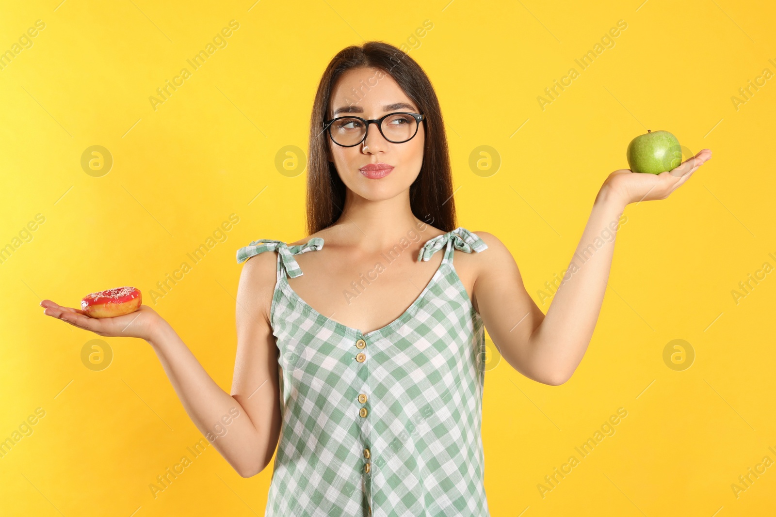 Photo of Doubtful woman choosing between apple and doughnut on yellow background