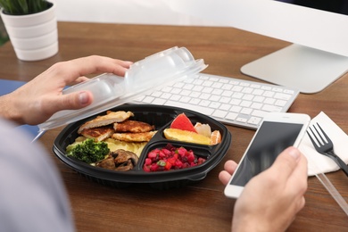 Office employee with smartphone having lunch at workplace, closeup. Food delivery