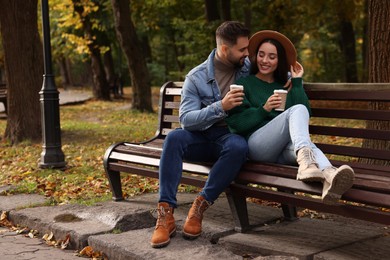 Photo of Happy young couple with cups of coffee spending time together on wooden bench in autumn park, space for text