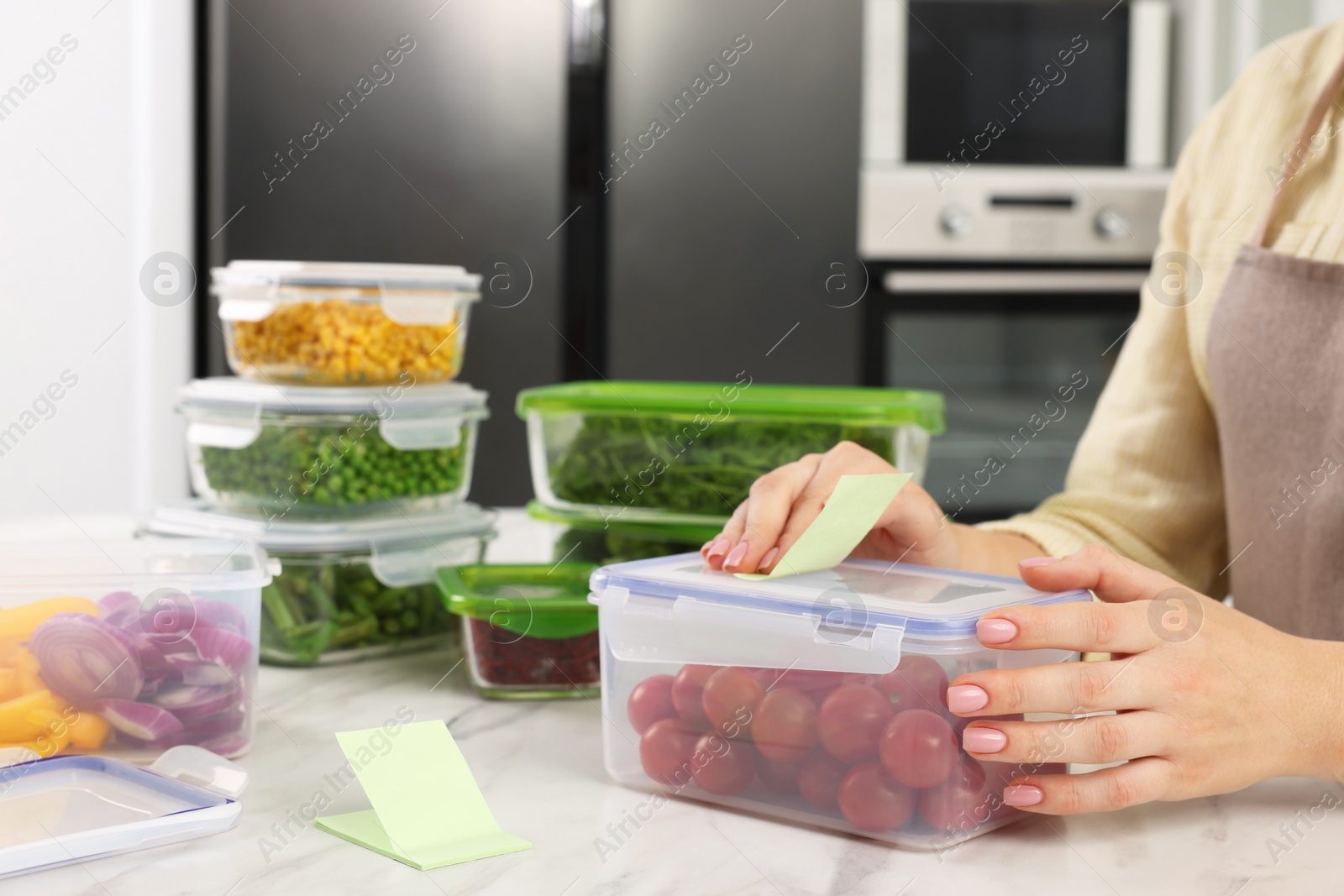 Photo of Woman sticking paper note onto container with fresh cherry tomatoes at white marble table in kitchen, closeup and space for text. Food storage