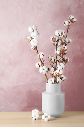 Beautiful cotton flowers in vase on wooden table against pink background