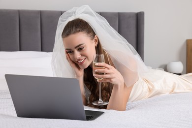 Happy bride with glass of sparkling wine and laptop on bed in bedroom