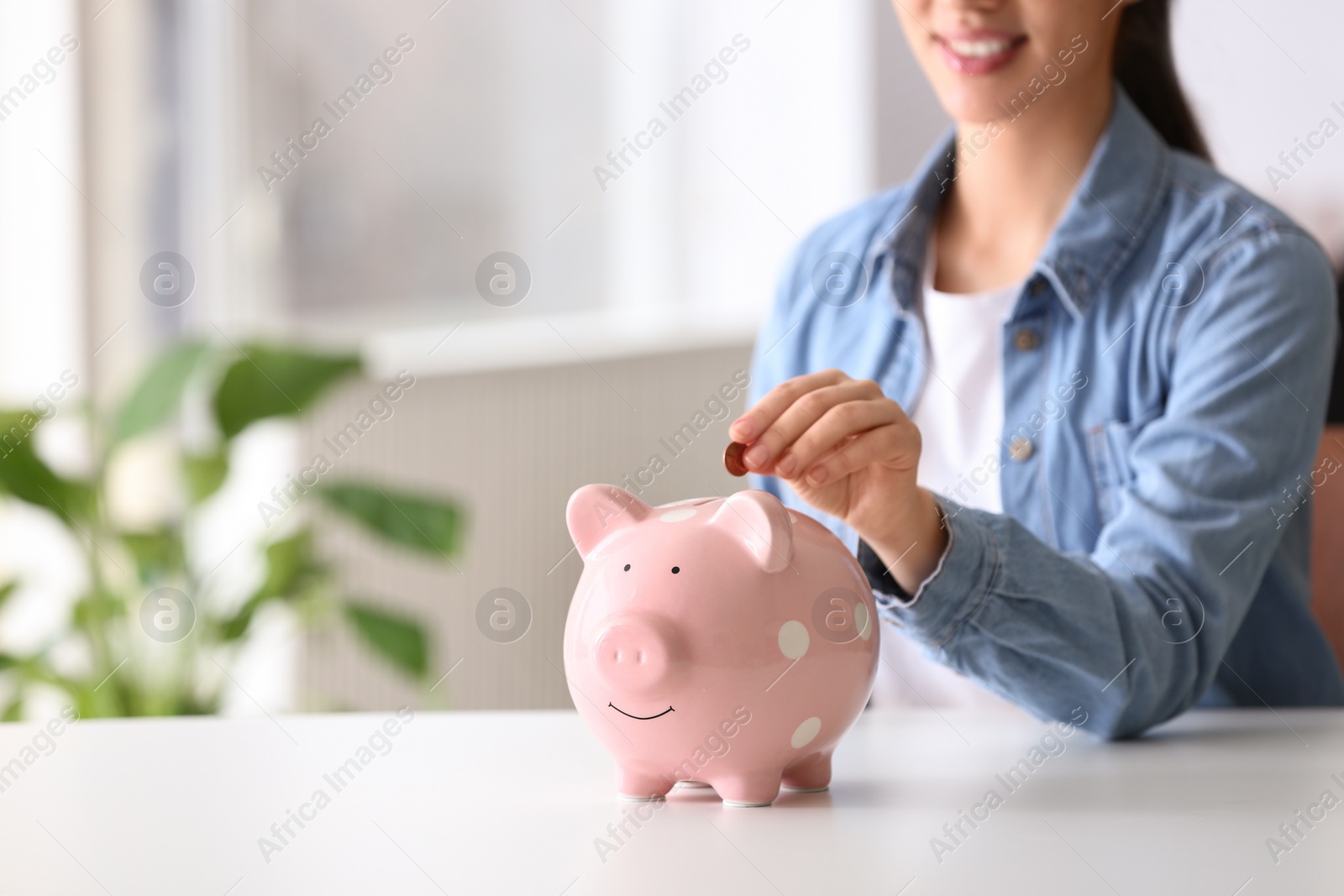 Photo of Woman putting coin into piggy bank at table indoors, closeup. Space for text