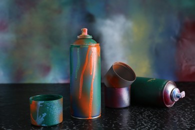 Two spray paint cans and caps on black textured surface against color background