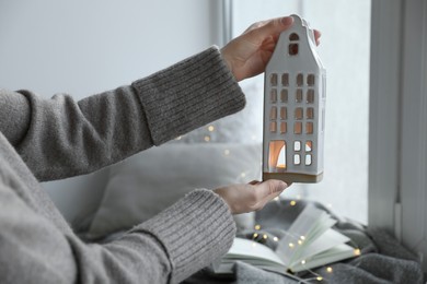 Photo of Woman with beautiful house shaped candle holder near window indoors, closeup