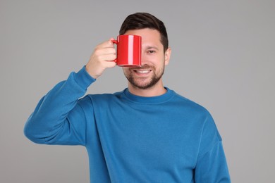 Portrait of happy man covering eye with red mug on grey background