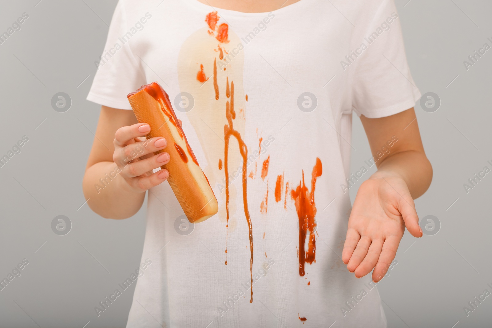 Photo of Woman holding hotdog and showing stain from sauce on her shirt against light grey background, closeup