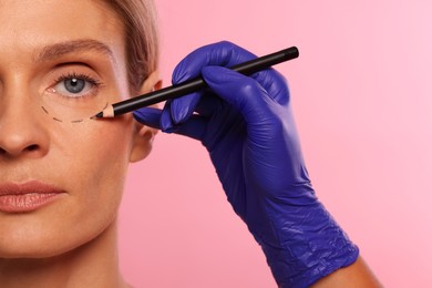 Photo of Doctor with pencil preparing patient for cosmetic surgery operation on pink background, closeup