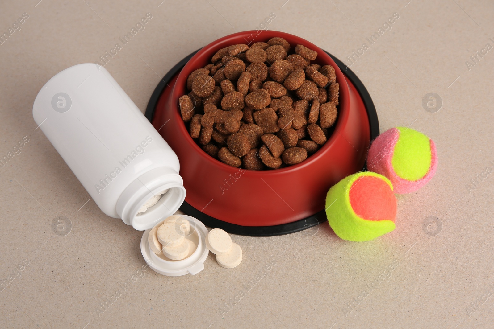 Photo of Bottle with vitamins, dry pet food in bowl and toy balls on beige background