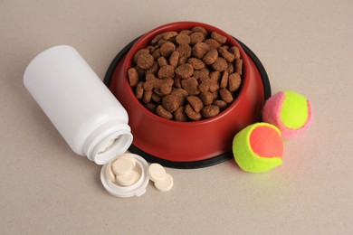 Bottle with vitamins, dry pet food in bowl and toy balls on beige background