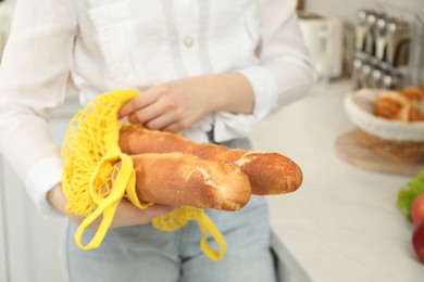 Photo of Woman holding string bag with baguettes in kitchen, closeup