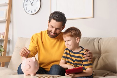 Cute little boy counting with calculator and his father putting coin into piggy bank at home