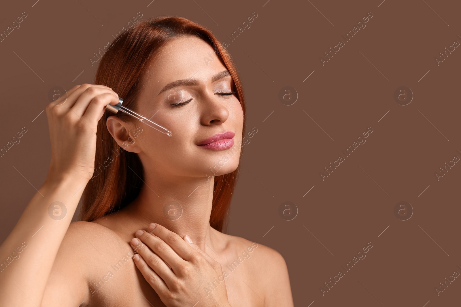 Photo of Beautiful young woman applying cosmetic serum onto her face on brown background, space for text