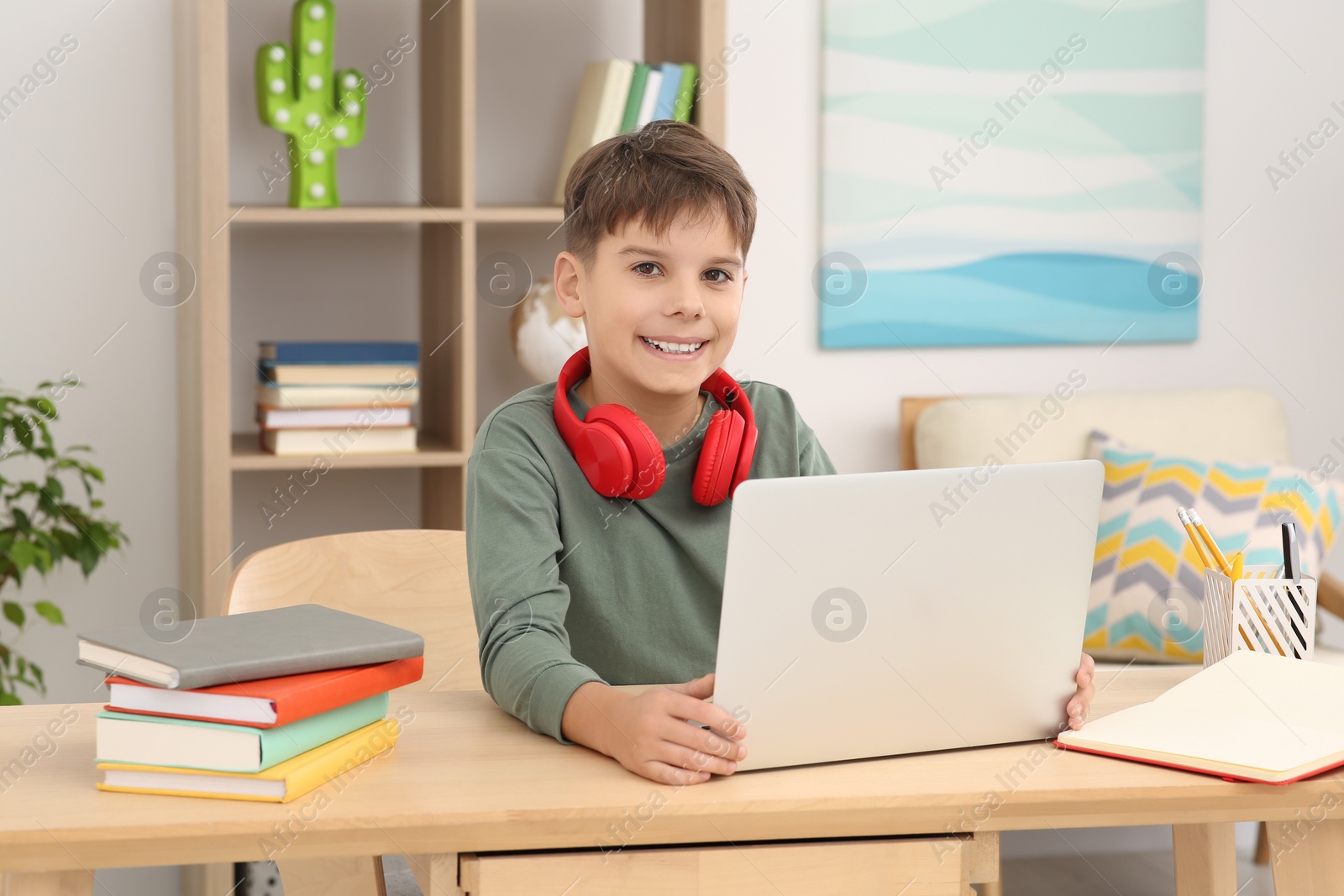 Photo of Boy with red headphones using laptop at desk in room. Home workplace