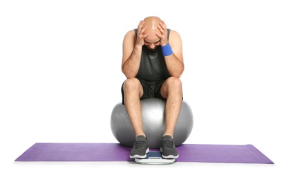 Upset overweight man with scale and fitness ball on white background