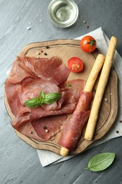 Photo of Delicious bresaola, tomato, grissini sticks and basil leaves on grey textured table, flat lay