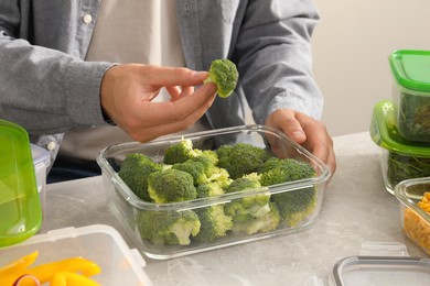 Man putting fresh broccoli into glass container at light grey table, closeup. Food storage