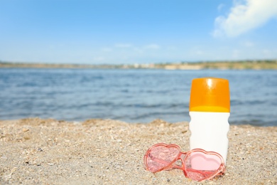 Photo of Bottle of sun protection body cream and glasses on beach, space for design