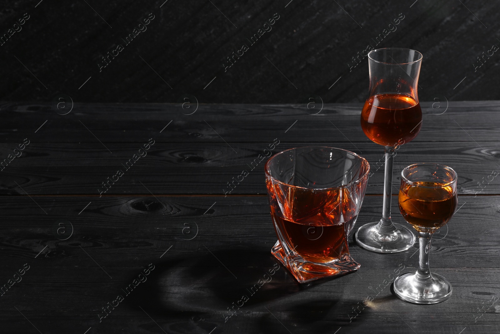 Photo of Different delicious liqueurs in glasses on black wooden table, space for text