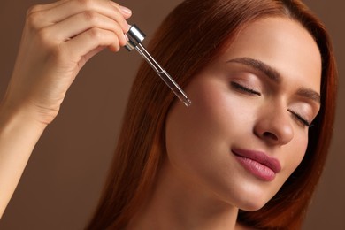 Beautiful young woman applying cosmetic serum onto her face on brown background, closeup