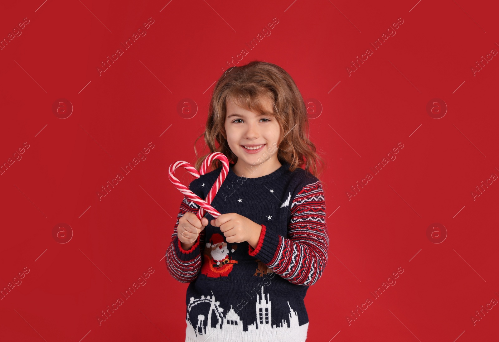 Photo of Cute little girl in Christmas sweater making heart shape with candy canes against red background