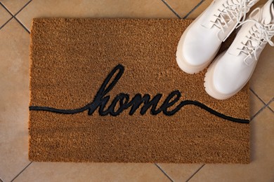 Photo of Doormat with word Home and stylish boots on tiled floor, flat lay
