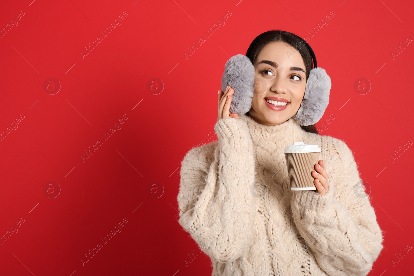 Photo of Beautiful young woman in earmuffs with cup of drink on red background. Space for text