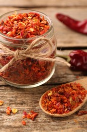 Chili pepper flakes in jar and spoon on wooden table, closeup