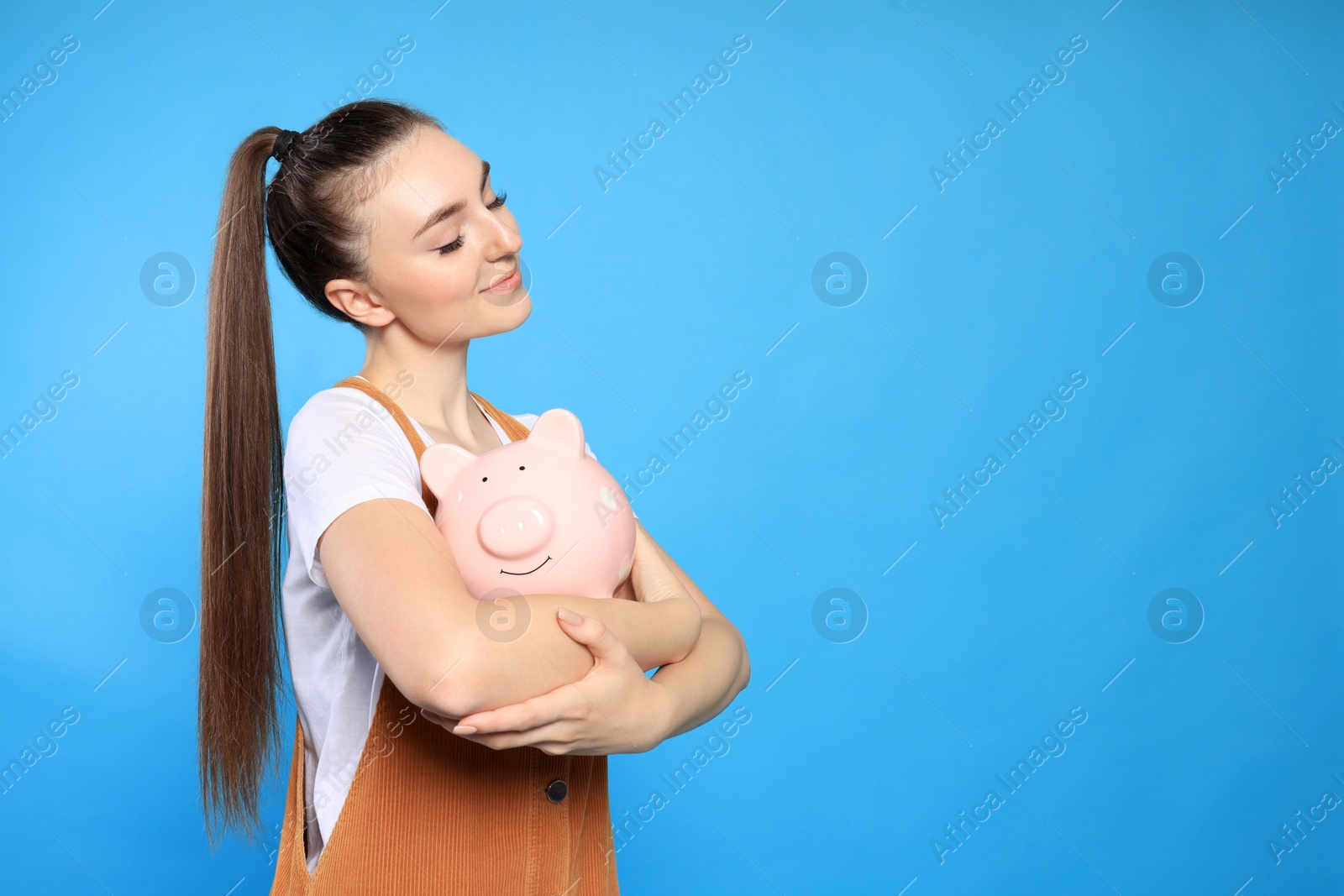 Photo of Happy young woman with piggy bank on light blue background. Space for text