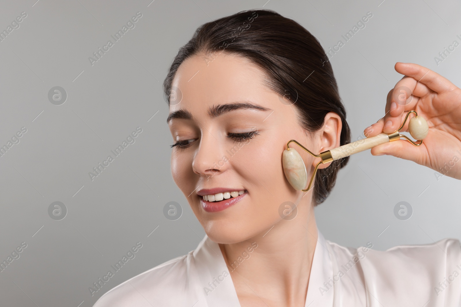 Photo of Young woman massaging her face with jade roller on grey background. Space for text