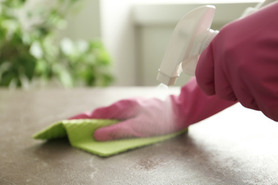 Photo of Woman in gloves cleaning brown marble table with spray detergent and rag indoors, closeup