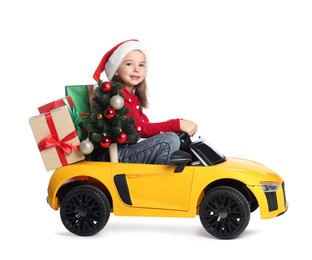 Photo of Cute little girl in Santa hat with Christmas tree and gift boxes driving children's electric toy car on white background