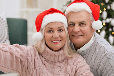 Happy mature couple in Santa hats taking selfie at home. Christmas celebration