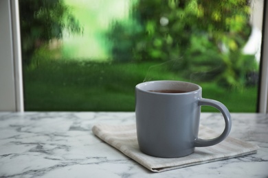 Photo of Cup of hot drink and napkin on marble windowsill against glass with rain drops, space for text